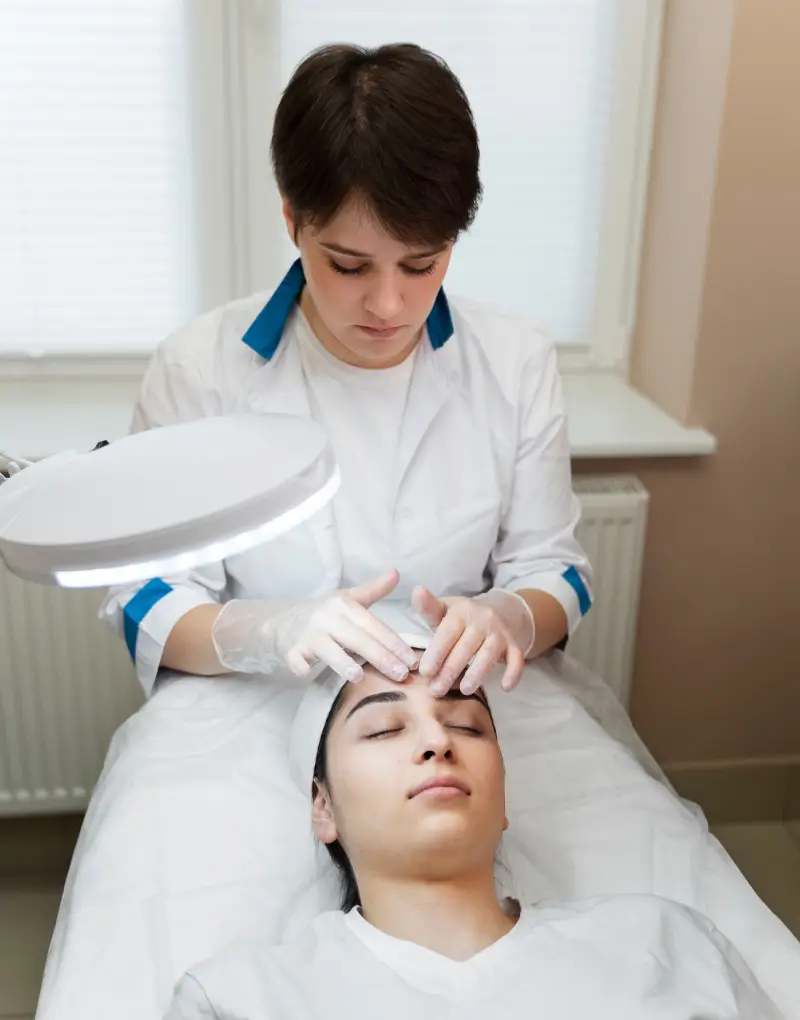 A healthcare professional performs a facial procedure under a bright lamp, while a patient lies on a treatment table